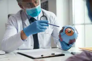 Ophthalmologist wearing personal protective equipment pointing to a model of an anatomical eyeball and showing it to a patient