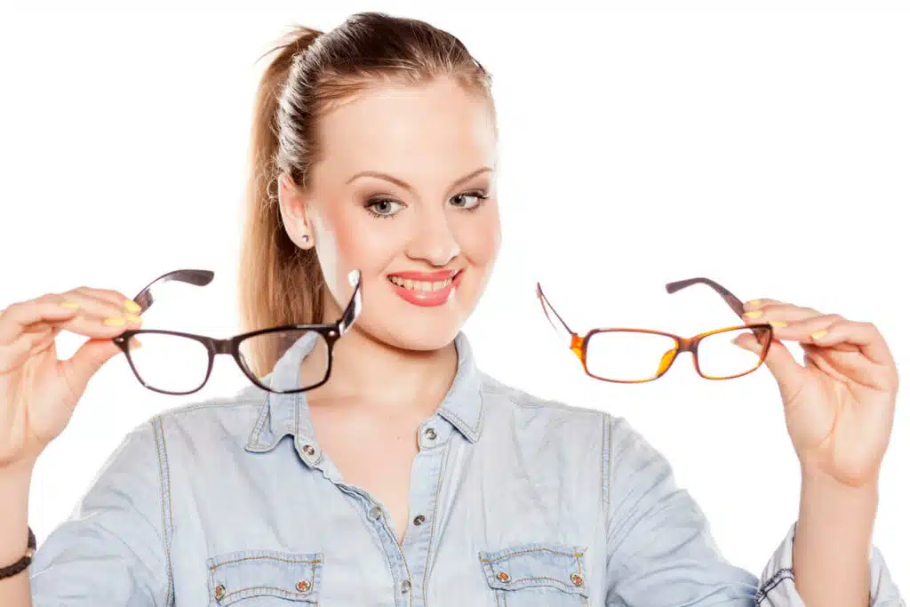 A young lady is smiling while holding a Same-Day Prescription Glasses and a Regular one, and trying to make a decision.